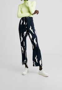 Marineblauwe wijde broek met wit abstract bladerenpatroon, voorzien van een strikceintuur. Gecombineerd met een aansluitende felgroene coltrui en witte sneakers.