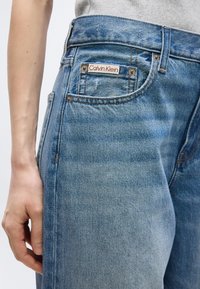 Κοντινό πλάνο ατόμου που φοράει μπλε τζιν Calvin Klein με εμφανές ετικέτα μάρκας στην μπροστινή τσέπη και γκρι μπλουζάκι τυλιγμένο μέσα.