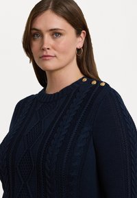 Marineblå strikkegenser med kabelmønstre, ribbet halslinning og tre gullknappeakenter på skulderen. Strukturert og chunky design.
