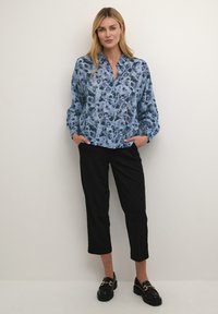 Camicia a bottone blu a fiori con maniche lunghe, indossata con pantaloni cropped neri e mocassini neri, caratterizzati da dettagli in oro.