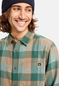 Camisa de franela de cuadros en verde y marrón con cierre de botones, un bolsillo en el pecho y un pequeño parche de logo negro en el lado izquierdo.