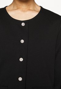 Černý oděv s kulatým výstřihem a pěti knoflíky s akcenty z krystalů. Vyrobeno z hladké látky se strukturovaným designem.