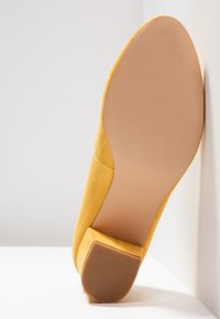 Chaussure plate en daim jaune avec une semelle lisse et un petit talon texturé, appuyée en angle contre un mur et une surface blancs.
