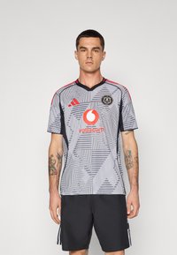Maglia sportiva grigia con un motivo geometrico, accenti neri e strisce rosse sulle spalle; presenta un collo rotondo e un logo sul petto.