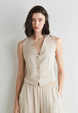 Jeune femme aux cheveux foncés mi-longs portant une veste sans manches beige boutonnée et un pantalon ample assorti, posant devant un mur uni.