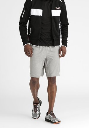 Mann trägt graue Shorts, schwarze Jacke mit weißen Streifen, schwarzes T-Shirt und graue Laufschuhe, geht vorwärts auf weißem Hintergrund.