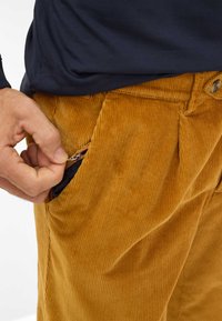 Katoenen broek in een warme bruine kleur met een gestructureerd oppervlak, voorzien van een ritszak aan de zijkant en geplooide ontwerpelementen.