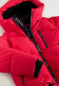 Chaqueta acolchada roja con capucha desmontable, con cierres de cremallera negros y un tirador de cremallera con logo. Tejido exterior suave con textura acolchada.