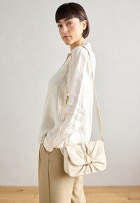 Beige leren handtas met een knoopdesign aan de voorkant, gouden hardware en een verstelbare schouderband, gedragen met een wit linnen shirt en beige broek.