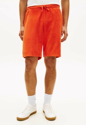 Homme portant un short orange vif jusqu'aux genoux, des chaussettes blanches et des baskets blanches, debout devant un fond blanc uni.