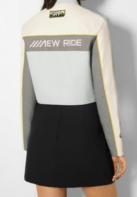 Veste en cuir bleu clair et gris avec des accents jaunes. Elle présente l'inscription "NEW RIDE" brodée au dos et un patch logo sur le col.