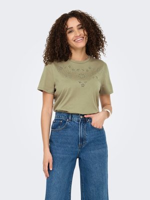 Donna sorridente con capelli ricci che indossa una maglietta verde oliva a maniche corte con ritagli floreali e jeans blu a vita alta, in piedi con una mano in tasca.