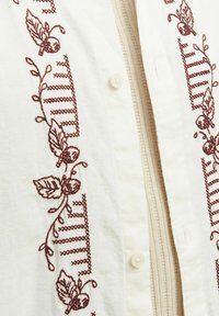 Broderet cremet skjorte med dybe bourgogne blomster- og geometriske mønstre. Har knaplukning og tekstureret stof.