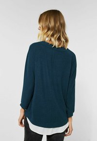 Kvinna med axellångt blont hår som bär en mörk teal långärmad tröja över en vit skjorta, vänd bort från kameran.