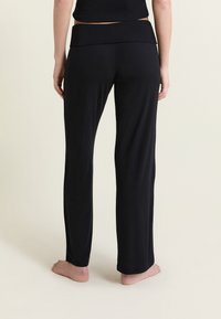 Vue de dos d'une personne portant un pantalon noir ample et un haut noir, debout pieds nus sur un sol clair avec un fond clair.