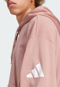 Hoodie rosa chiaro con una texture morbida, caratterizzato da un logo Adidas bianco sulla manica. Include un cappuccio con coulisse e polsini a coste.