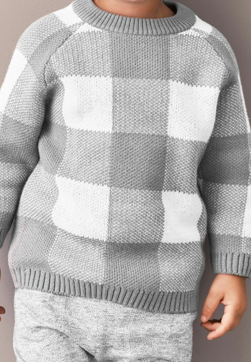 Enfant portant un pull en tricot à motif à carreaux gris et blanc et un pantalon gris clair, debout devant un fond beige.
