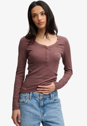 Jeune femme aux cheveux foncés portant une chemise marron à manches longues boutonnée et un jean bleu clair, posant avec une main sur la taille.