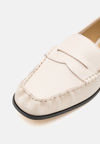 Mocassin en cuir couleur crème avec détails cousus et semelle noire, présenté en gros plan sur un fond blanc.