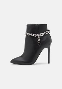 Botín negro de tobillo hecho de cuero suave con punta afilada y tacón stiletto de 9 cm, con un detalle de cadena plateada alrededor del tobillo.