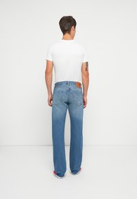 Homem de costas, vestindo uma t-shirt branca, jeans de perna reta azuis, ténis vermelhos estilo Converse, e uma tatuagem no braço esquerdo.