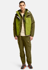 Chaqueta verde con cremalleras y bolsillos, superpuesta sobre una blusa beige. Combinada con pantalones cargo verdes y zapatos de senderismo negro y naranja.