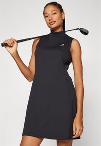 Μαύρο αμάνικο γκολφ φόρεμα με ψηλό λαιμό, φτιαγμένο από εύκαμπτο ύφασμα. Έχει ένα έμβλημα στο στήθος και πλαϊνές τσέπες.