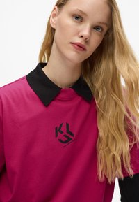 Blondínka s dlhými vlasmi, oblečená v magentovom tričku s čiernym golierom a čiernym textom loga "Karl Lagerfeld Jeans" na hrudi.
