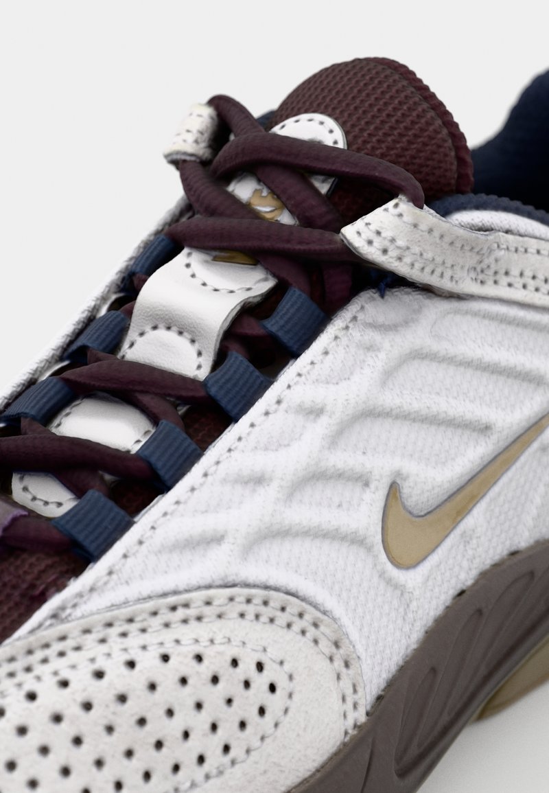 Sneaker Nike présentant une tige en maille blanche avec des accents bordeaux et marine, des superpositions texturées, des lacets plats et un détail logo doré.