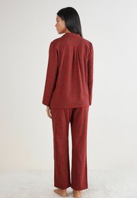 Rødternet pyjamassæt bestående af en langærmet blazer med knaplukning og løse bukser, lavet af blødt stof med en tekstureret overflade.