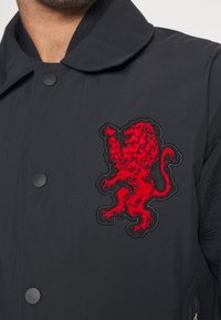 Manteau noir avec un emblème rouge de lion bien visible ; tissu lisse avec fermeture à boutons et col plat, mettant en valeur un design épuré.