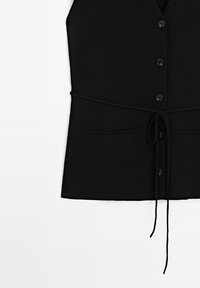 Svart stickad väst med V-ringning, fem knappar framtill och ett midjeband. Har två fickor framtill och en slät textur.