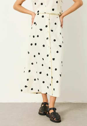 Frau trägt einen cremefarbenen, knöpfigen Midirock mit schwarzen Punkten und schwarzen, geflochtenen, geschlossenen Sandalen, steht mit den Händen in den Hüften.