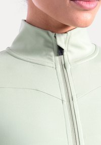 Chaqueta verde claro con cremallera, cuello alto, costuras planas y cremallera negra. Tejido liso con detalles sutilmente texturizados.