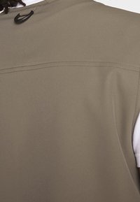 Khaki ärmlös väst med en slät textur, vertikal sömdetalj och en liten ögla för upph�ängning på baksidan. Vit undertröja synlig under.