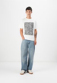 Hvit bomull t-skjorte med svart grafisk design, parret med løstsittende lyseblå denimjeans og hvite joggesko.