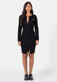 Vrouw met lang golvend haar, gekleed in een aansluitende zwarte blousejurk en zwarte loafers, staande tegen een effen lichte achtergrond.