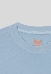 T-shirt en coton bleu clair avec un col rond. L'étiquette à l'intérieur affiche un texte orange avec un logo d'aile et le nom de la marque "KARORAL."