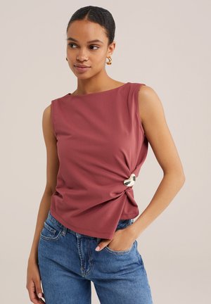 Donna che indossa un top senza maniche color malva con dettaglio asimmetrico di chiusura in argento sul lato, abbinato a jeans blu, guarda leggermente di lato.