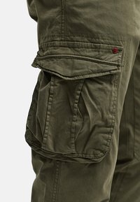 Pantalones cargo en algodón verde oliva, con grandes bolsillos laterales, detalles de costura visibles y un pequeño botón de acento rojo.