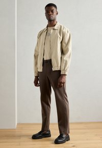 Ein Mann steht und trägt eine beige Reißverschlussjacke, ein hellbeiges Hemd mit Grafik, braune Hose und schwarze Mokassins auf einem Holzboden vor einer schlichten Wand.