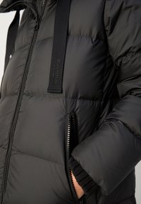 Schwarze Daunenjacke mit glatter, glänzender Oberfläche, ausgestattet mit Reißverschluss-Taschen und elastischen Bündchen. Enthält ein Logo am Schulterriemen.