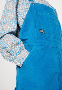 Jasnoniebieskie ogrodniczki z sztruksu noszone na szarej koszuli z niebiesko-białymi geometrycznymi wzorami, ręka w bocznej kieszeni.