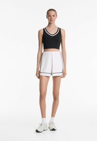 Sort cropped tanktop med hvide striber, parret med hvide shorts med sorte accenter og folder; modellen har hvide sneakers og sokker på.