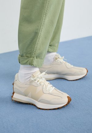Person iført beige sneakers med hvide snørebånd, hvide sokker og op rullede lysegrønne bukser, står på en blå gulv.