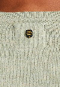 Hellgrüner Strickpullover mit einem gerippten Kragen. Verziert mit einem schwarzen Etikett mit "PME LEGEND" in Gelb, das mit einer braunen Schnur befestigt ist.