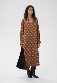 Rjava obleka z dolgimi rokavi in V-izrezom, okrašena z belimi pikami in nabranimi zvončastimi rokavi. Oblečena skupaj s črno, strukturno torbo tote.