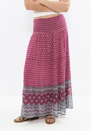 Femme portant une longue jupe rose magenta à motifs avec une large ceinture, assortie à des sandales marron cloutées, debout les bras croisés.