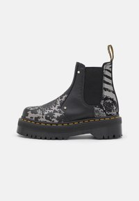 Black Chelsea boots featuring a contrasting gray patterned side, textured black elastic panels, and yellow stitching on a thick rubber sole.