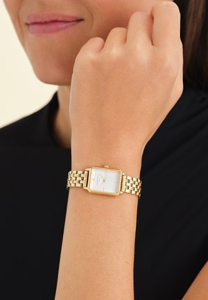 Montre-bracelet dorée avec un cadran blanc rectangulaire, un bracelet en métal maillé et des marqueurs d'heure simples sur le cadran. Peau visible sur un poignet.